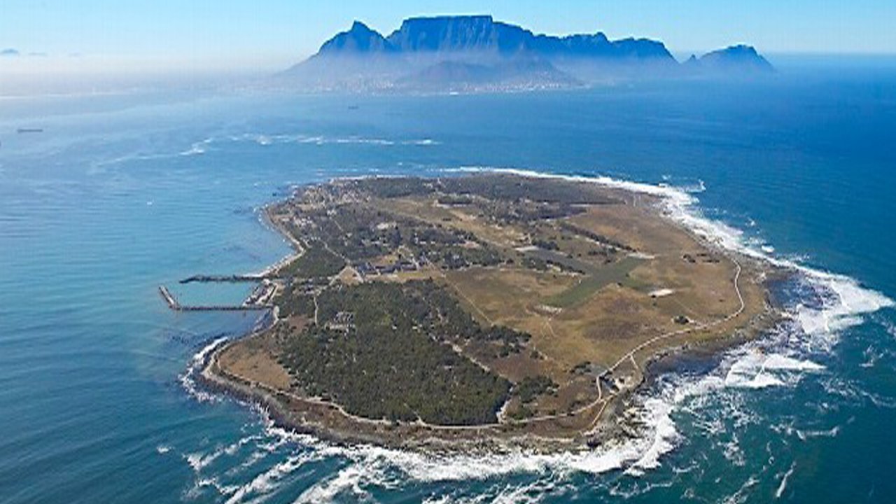 Aerial view of a small island with a mountain range in the background across the ocean; blue sea and sky with green and earthy tones